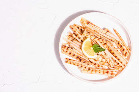 Grilled scorzonera, trendy vegan food. Modern hard light, dark shadow. Ceramic stand, lemon, parsley. White plaster background, top viewの写真素材