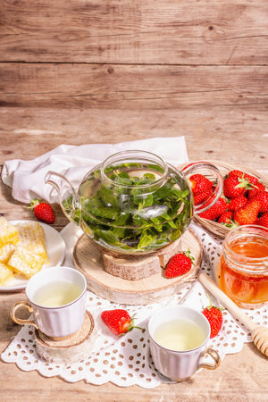 Teatime concept. Aromatic mint tea, ripe strawberries, sweet honey. A hot drink, ceramic cups on stands, vintage linen napkin. Old wooden boards background, copy spaceの写真素材