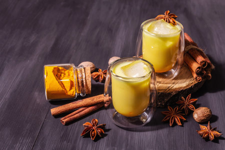 Golden turmeric milk with ice. Cinnamon, nutmeg, anise spices. Trendy healthy drink concept. A modern hard light, dark shadow, black wooden background, copy spaceの写真素材