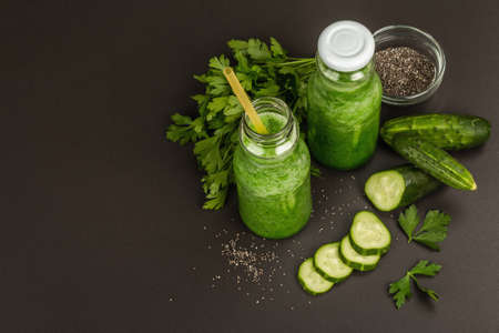 Green smoothie with cucumber in glass bottles. Fresh ripe vegetables, greens, and chia seeds. Black stone concrete background, top viewの写真素材