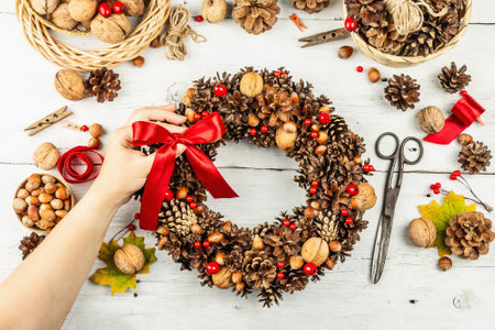 Female hands hold a tied bow for an autumn wreath of cones, nuts, and berries. Natural materials for creative composition. Handmade home decoration festive concept. White wooden background, top viewの写真素材