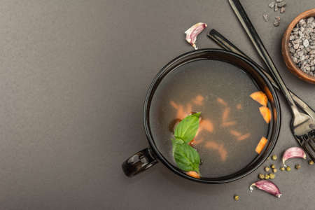 Transparent chicken broth with vegetables in a cup. Traditional bouillon, healthy food, cutlery. Black stone concrete background, top viewの写真素材