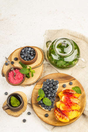 Tasty toast bread with cream cheese and ripe fruits. Grilled peach, blueberries, and mint tea on the...