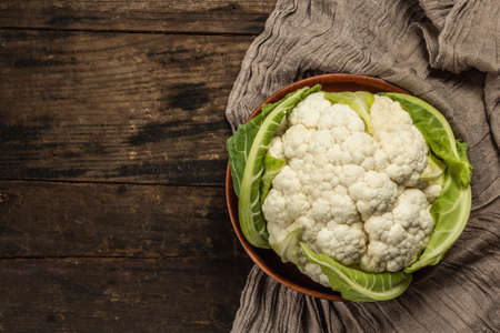 Beautiful cauliflower with green leaves in a ceramic bowl. Vintage napkin, old wooden table, top viewの写真素材