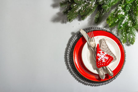 Christmas table setting. Red ceramic plate with vintage cutlery, fir tree branches, and festive accessories. Modern hard light, dark shadow. Creative flat lay, gray background, top viewの写真素材