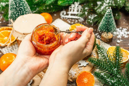 Woman's hands hold orange marmalade or orange jam in a glass jar. Sweet confiture with festive Christmas decor. Traditional New Year food, wooden background, copy spaceの写真素材