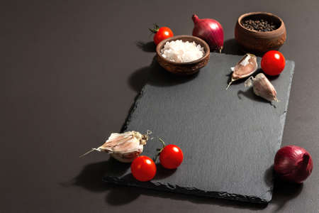 Cooking concept. Ceramic stand, tomato cherries, garlic, sea salt, pepper, and purple onion. A trendy hard light, dark shadow, black stone concrete background, copy spaceの写真素材