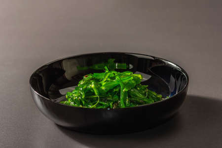 Wakame seaweed salad with sesame seeds and chili pepper in a bowl. Trendy hard light, dark shadow. Black stone concrete background, flat lay, copy spaceの写真素材