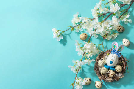 Happy Easter blue background with blooming cherry, quail eggs in a nest and cute rabbit. Spring flat...