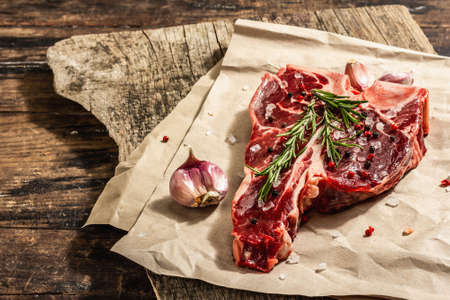 Classic T-bone steak with fresh rosemary, garlic and spices. A modern hard light, dark shadow, trendy stand, wooden background, flat lay. Wooden background, rustic style, copy spaceの写真素材
