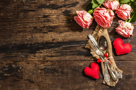 Rustic table setting for romantic dinner. Bouquet of fresh roses, cutlery and heart. Vintage wooden board background, top viewの写真素材