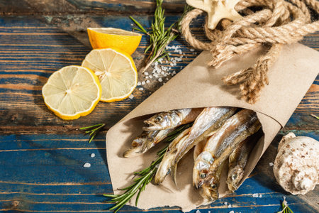 Sun-dried smelt in a paper package. Salted fish with marine decor. Lemon, fresh rosemary, sea rope. Blue nautical wooden background, top viewの写真素材