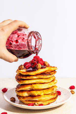 Classic homemade American pancakes with frozen raspberries, hazelnut and mint. The male hand holds a jar of fruit jam. Stone concrete background. Trendy hard light, dark shadow, place for textの写真素材