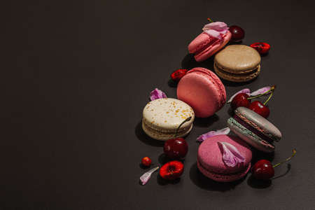 Macarons with peonies flower petals and ripe cherries on a black stone background. Sweet dessert, romantic style, festive card, flat lay, hard light, dark shadow, copy spaceの写真素材