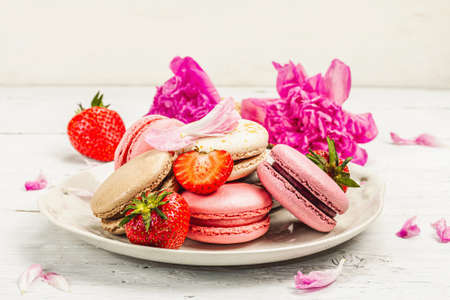 Macarons with strawberries and peonies flower petals, on a white wooden background. Sweet dessert, romantic style, flat lay, hard light, dark shadow, close upの写真素材