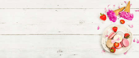 Macarons with strawberries and peonies flower petals, on a white wooden background. Sweet dessert, romantic style, flat lay, hard light, dark shadow, banner formatの写真素材
