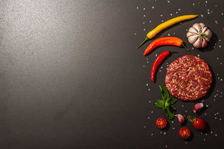 Raw steak cutlets from minced beef meat on black stone background. Fresh vegetables, mint, and...
