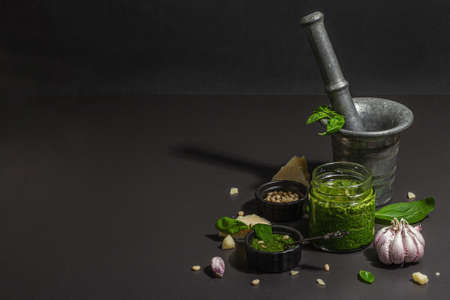 Preparation process Italian basil pesto sauce in a vintage mortar with a pestle. Homemade food, ingredients for a traditional recipe. Hard light, dark shadow, black stone background, copy spaceの写真素材