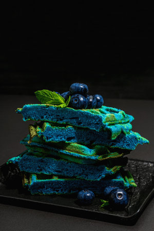 Homemade Belgium Waffles with blueberries. Gluten-free blue dessert, fresh fruits and mint. Trendy hard light, dark shadow. Black stone concrete background, copy spaceの写真素材