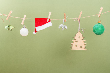 Festive New Year background. Traditional symbols hanging on the thread: fir tree, Christmas balls, cones, Santa cap, bells. Trendy Savannah Green color backdrop, place for textの写真素材