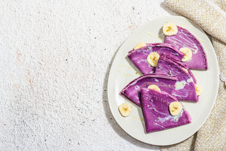 Thin lavender pancakes with banana slices and sweet toppings. Healthy vegan non lactose and gluten free food, breakfast concept. Trendy hard light, dark shadow, light plaster background, top viewの写真素材