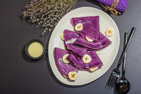 Thin lavender pancakes with banana slices and sweet toppings. Healthy vegan non lactose and gluten free food, breakfast concept. Trendy hard light, dark shadow, black stone background, top viewの写真素材