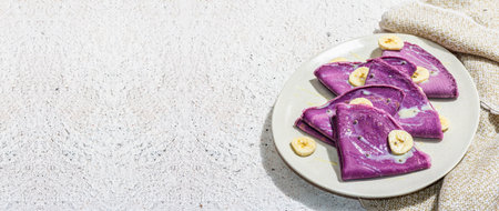 Thin lavender pancakes with banana slices and sweet toppings. Healthy vegan non lactose and gluten free food, breakfast concept. Trendy hard light, dark shadow, light plaster background, banner formatの写真素材