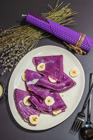 Thin lavender pancakes with banana slices and sweet toppings. Healthy vegan non lactose and gluten free food, breakfast concept. Trendy hard light, dark shadow, black stone background, top viewの写真素材