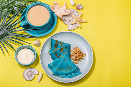 A cup of coffee and blue thin pancakes in a marine style. Starfish, sea shells, palm leaves. Hard light, dark shadow, bright yellow background, top viewの写真素材