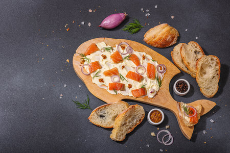 Butter Board food trend topped with salmon, shallots and fresh greens. Appetizer for a party, baguette pieces, hard light, dark shadow. Black stone concrete background, flat lay, top viewの写真素材