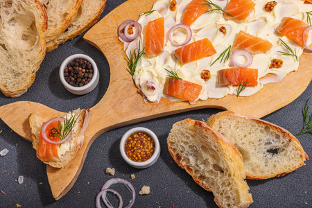 Butter Board food trend topped with salmon, shallots and fresh greens. Appetizer for a party, baguette pieces, hard light, dark shadow. Black stone concrete background, flat lay, top viewの写真素材
