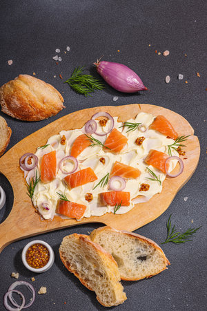 Butter Board food trend topped with salmon, shallots and fresh greens. Appetizer for a party, baguette pieces, hard light, dark shadow. Black stone concrete background, flat lay, top viewの写真素材