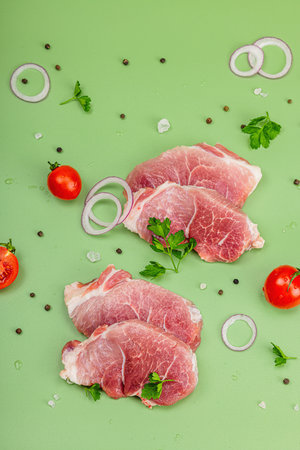 Pieces of pork meat with parsley, purple onion rings and tomato, raw fresh steaks concept. Sea salt, spices, spring green background, water drops, flat lay, top viewの写真素材