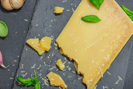 Piece of Parmesan cheese on modern stand. Garlic, fresh basil leaves, traditional Italian ingredients for healthy eating. Black stone concrete background, flat lay, top viewの写真素材