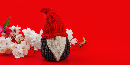 Minimalist spring greeting card with knitted gnome and blooming cherry branches. Traditional festive symbol, handmade concept. Passion red background, banner formatの写真素材