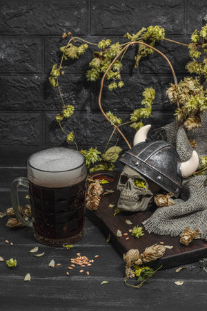 Creative Oktoberfest composition. Dark beer, viking helmet, hop and wheat. German festival food concept. Trendy hard light, dark shadow, vintage black background, close upの写真素材