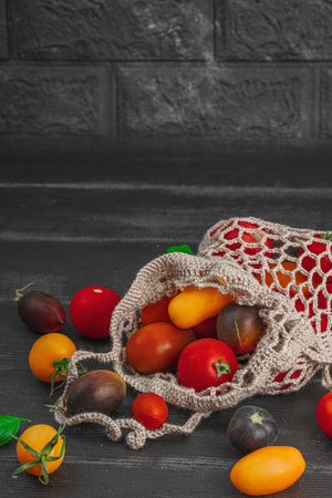 Assortment of various ripe tomatoes in a trendy eco bag. Handmade, crocheted, zero-waste concept. Black wooden background, copy spaceの写真素材