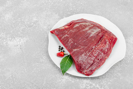 Fresh raw flank steak with assorted spices on a white ceramic plate. Light stone background. Culinary concept for meat preparation, gourmet cooking, or food styling, copy spaceの写真素材