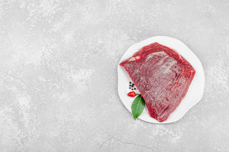 Fresh raw flank steak with assorted spices on a white ceramic plate. Light stone background. Culinary concept for meat preparation, gourmet cooking, or food styling, top viewの写真素材