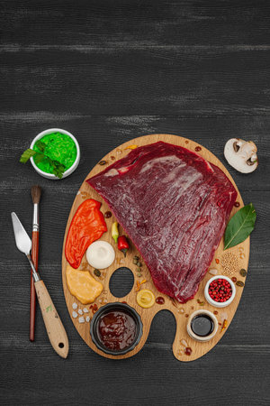 Gourmet food concept with raw flank steak, assorted spices, sauces, and herbs arranged creatively on wooden painter's palette. Chef artistry and culinary creativity, black wooden background, top viewの写真素材
