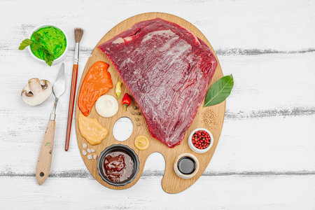 Gourmet food concept with raw flank steak, assorted spices, sauces, and herbs arranged creatively on wooden painter's palette. Chef artistry and culinary creativity, white wooden background, top viewの写真素材