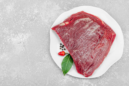 Fresh raw flank steak with assorted spices on a white ceramic plate. Light stone background. Culinary concept for meat preparation, gourmet cooking, or food styling, top viewの写真素材