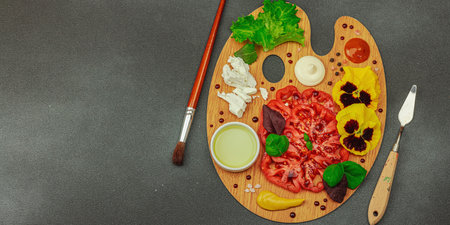 Sliced heirloom tomato with feta, herbs, edible flowers, and sauces on a wooden artist palette, styled with brush and palette knife. Dark stone concrete background, banner formatの写真素材