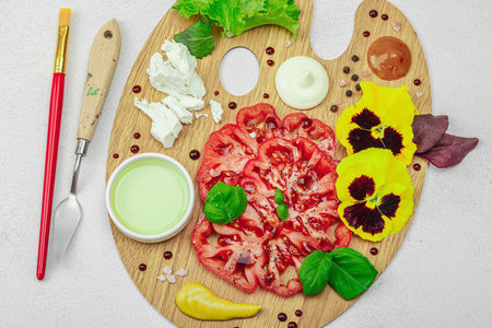 Sliced heirloom tomato with feta, herbs, edible flowers, and sauces on a wooden artist palette, styled with brush and palette knife. Light stone concrete background, top viewの写真素材