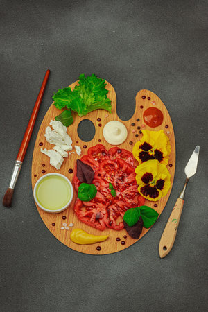 Sliced heirloom tomato with feta, herbs, edible flowers, and sauces on a wooden artist palette, styled with brush and palette knife. Dark stone concrete background, top viewの写真素材