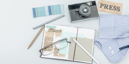 Flat lay of retro journalist essentials on blue background: camera, press badge, notebook, newspaper...