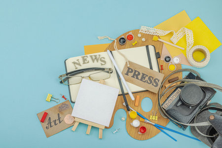 Flat lay of press badge, vintage camera, art supplies, notebook, and exhibition label on a blue...