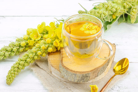 Glass cup of mullein herbal tea on natural wooden background with fresh yellow Verbascum thapsus...