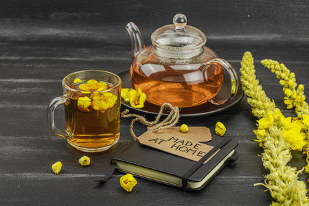 Glass cup of mullein flower tea with teapot and notebook on black wooden table, healthy natural...