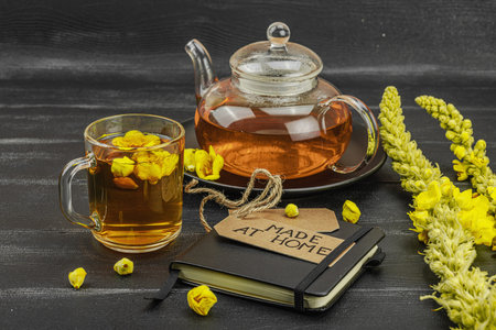 Glass cup of mullein flower tea with teapot and notebook on black wooden table, healthy natural...
